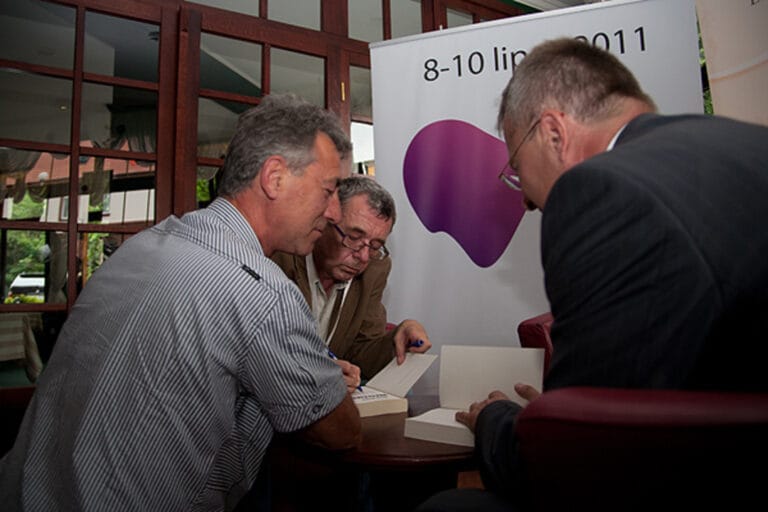 Mężczyźni podpisują książki obok plakatu konferencji.