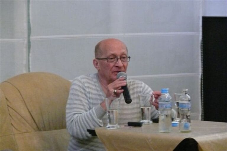 Mężczyzna mówi do mikrofonu przy stoliku z wodą.