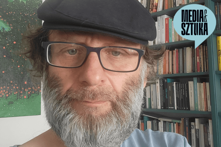 Mężczyzna w czapce z brodą przed półką z książkami.