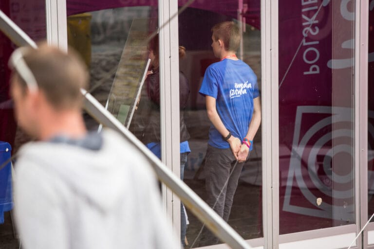 Osoba w niebieskiej koszulce przed szybą.