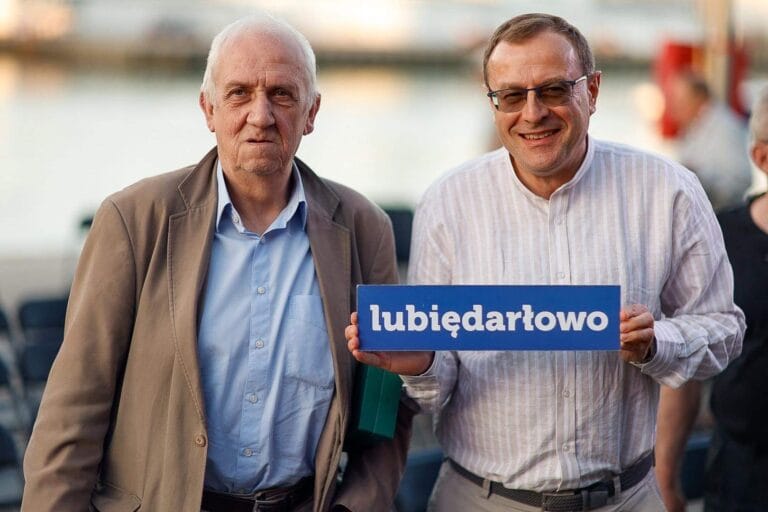 Dwóch mężczyzn z tabliczką 'lubiędarłowo' nad wodą.