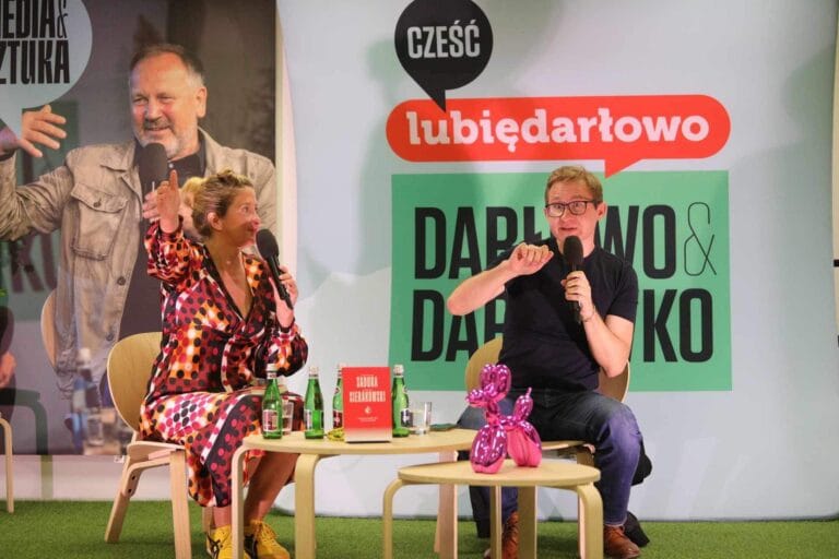 Dwóch ludzi prowadzi rozmowę na scenie z plakatem.