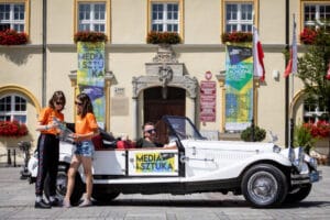 Ludzie przy zabytkowym samochodzie na tle ratusza.