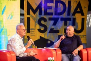 Wywiad na kanapie, mikrofony w dłoniach, plakat w tle.