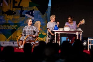 Aktorzy występujący na scenie, siedzący przy stole.