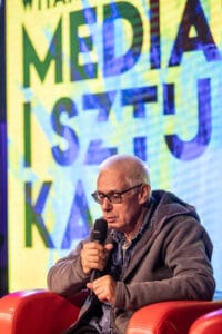 Mężczyzna na scenie z mikrofonem, napis 'MEDIA' w tle.