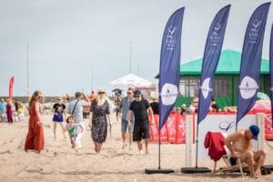 Ludzie spacerują po plaży przy flagach i parasolach.