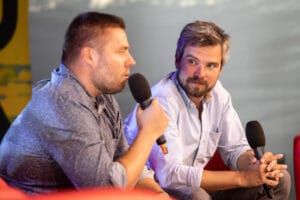 Dwóch mężczyzn z mikrofonami na scenie, rozmowa.