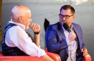 Dwaj mężczyźni dyskutują na scenie, mikrofon w ręku.