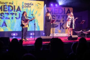 Trzy osoby występują na scenie z mikrofonami.