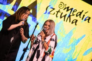 Dwie kobiety śpiewają na scenie przed kolorowym tłem.